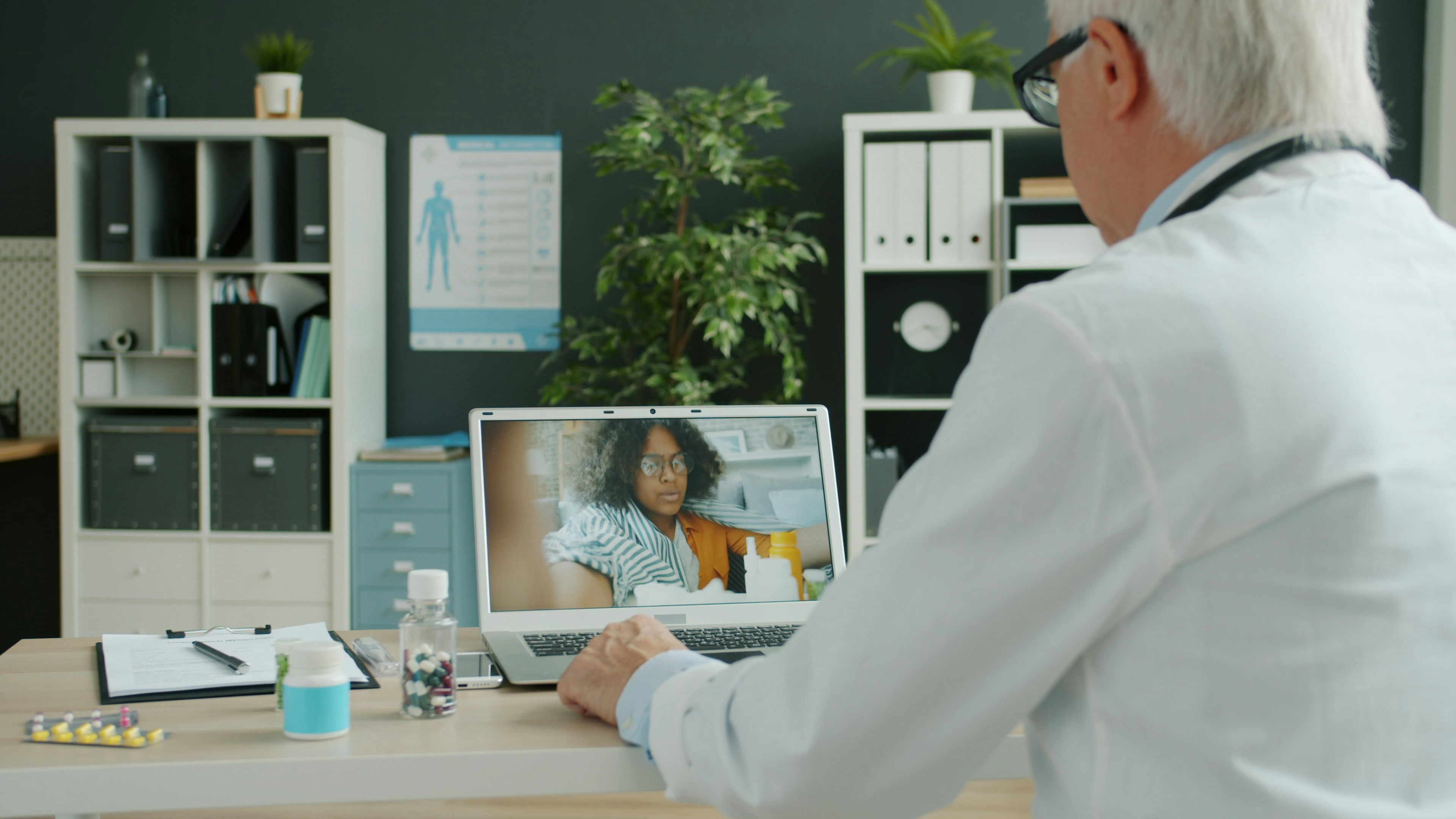 Senior doctor in white gown is consulting young Afro-American woman online discussing treatment and pills working from hospital using computer.